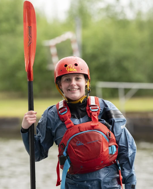 Smiling young woman in a red helmet and buoyancy aid holding a kayak paddle by the water. Smiling young woman in a red helmet and buoyancy aid holding a kayak paddle by the water.